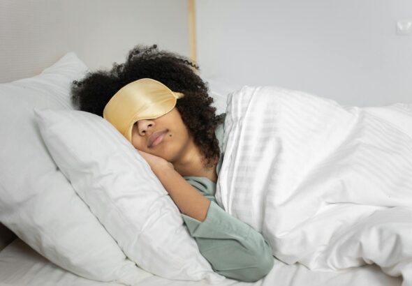 A woman with afro hair sleeps soundly in bed with a sleep mask, enjoying a cozy indoor atmosphere.